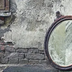 Owalne, stare lustro w drewnianej ramie opiera się o zniszczoną, ceglaną ścianę.